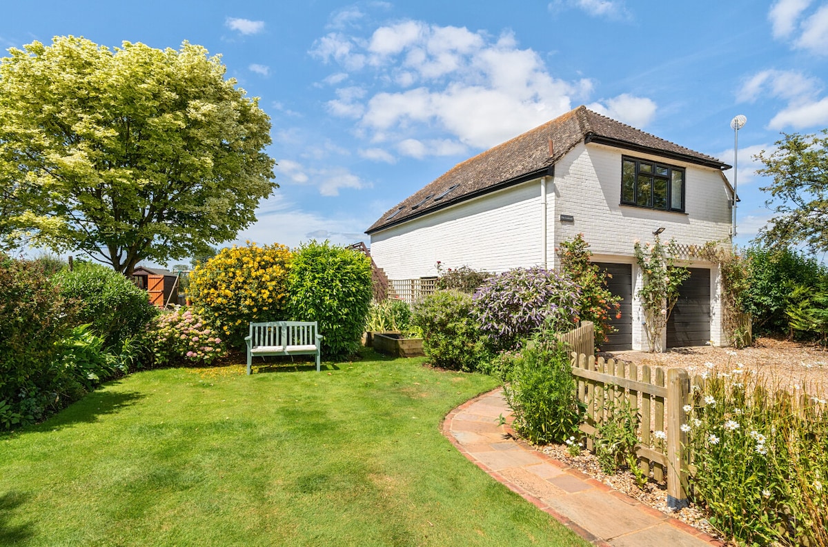 A spacious garden surrounds a charming two-story building, featuring white walls and a dark roof. Lush greenery and flowering shrubs add color to the landscape. A wooden bench is positioned on the lawn, inviting moments of relaxation amidst the tranquil setting.