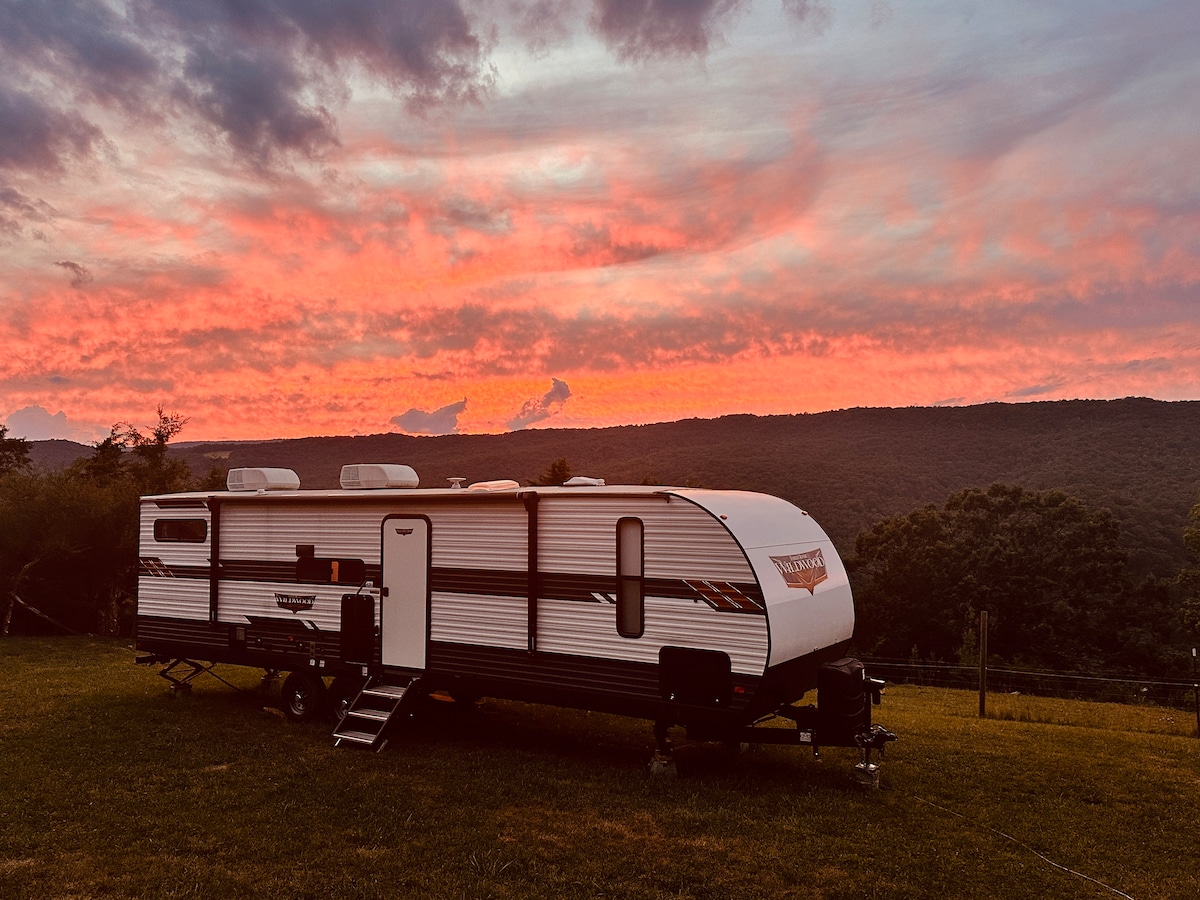 A charming camper is positioned on an expansive grassy area, set against a vibrant sunset sky filled with shades of orange and pink. The surrounding mountains provide a serene backdrop, enhancing the sense of peace in the landscape.