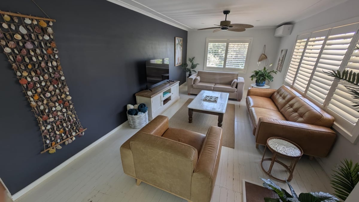 A spacious living area features two leather sofas and a fabric couch arranged around a wooden coffee table. A television is mounted on the wall, complemented by decorative artwork. Plants are placed throughout, and natural light filters through the window covered by shutters.