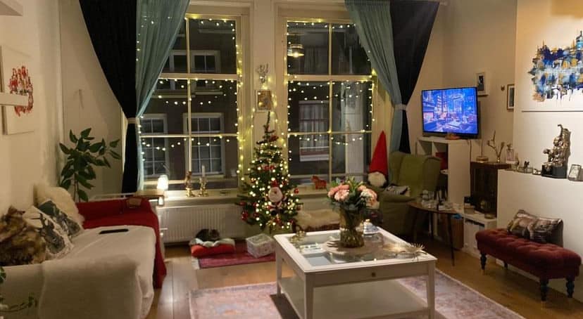 A cozy living area is illuminated by soft string lights, creating a warm ambiance. A Christmas tree adorned with lights stands in the corner. A coffee table is centrally positioned, surrounded by comfortable seating, while art pieces add character to the walls.