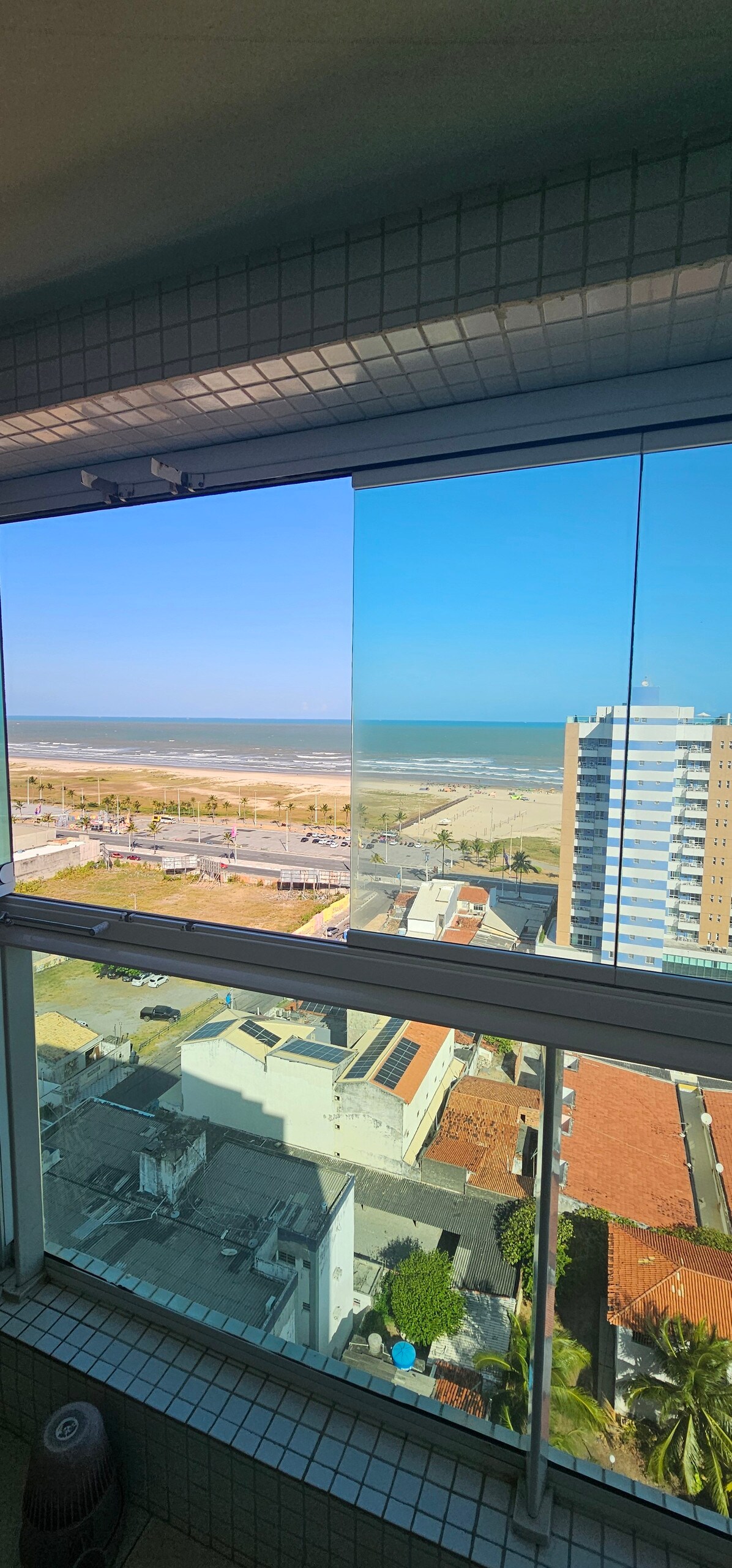 A panoramic view of the coastline is captured through glass sliding doors, revealing a clear blue sky and the ocean. The expansive beach stretches below, framed by buildings and greenery, enhancing the connection to the seaside environment.