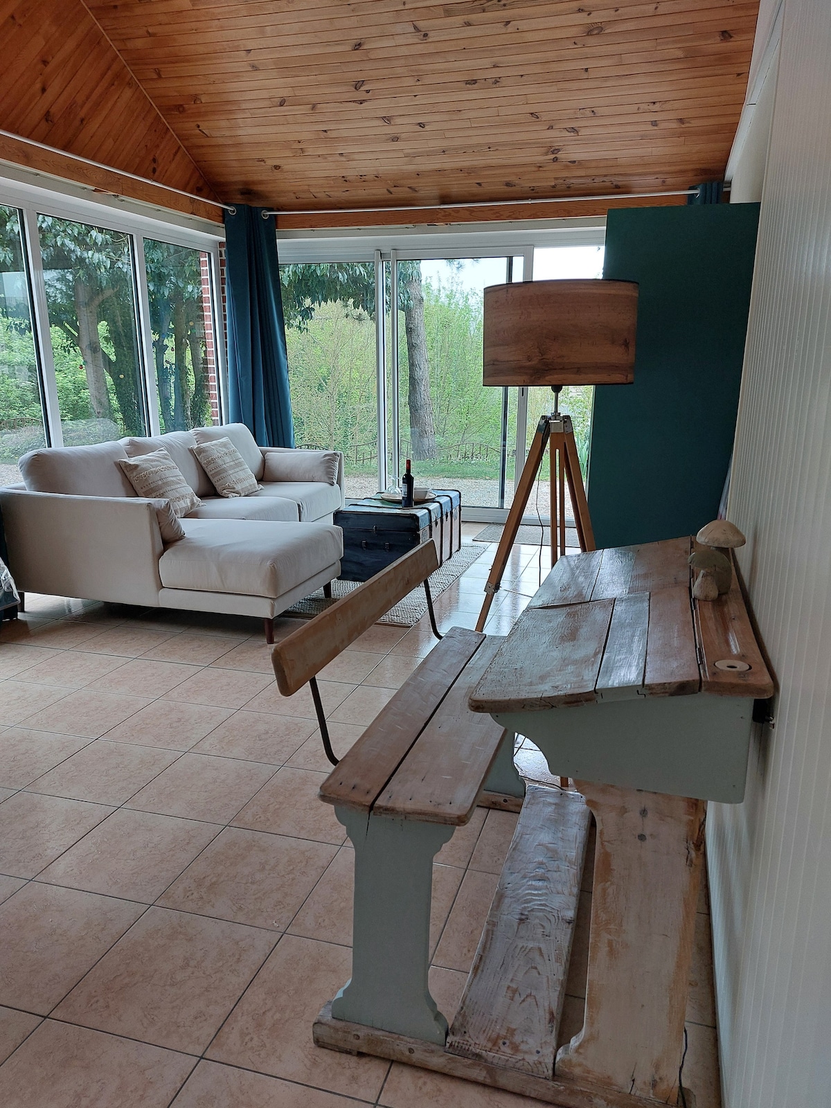 A sunlit veranda features a light-colored sofa with cushions, positioned beside a tall lamp. A rustic wooden table stands nearby, with a view of greenery through large glass doors. Natural wood beams create a warm ceiling overhead, enhancing the inviting space.