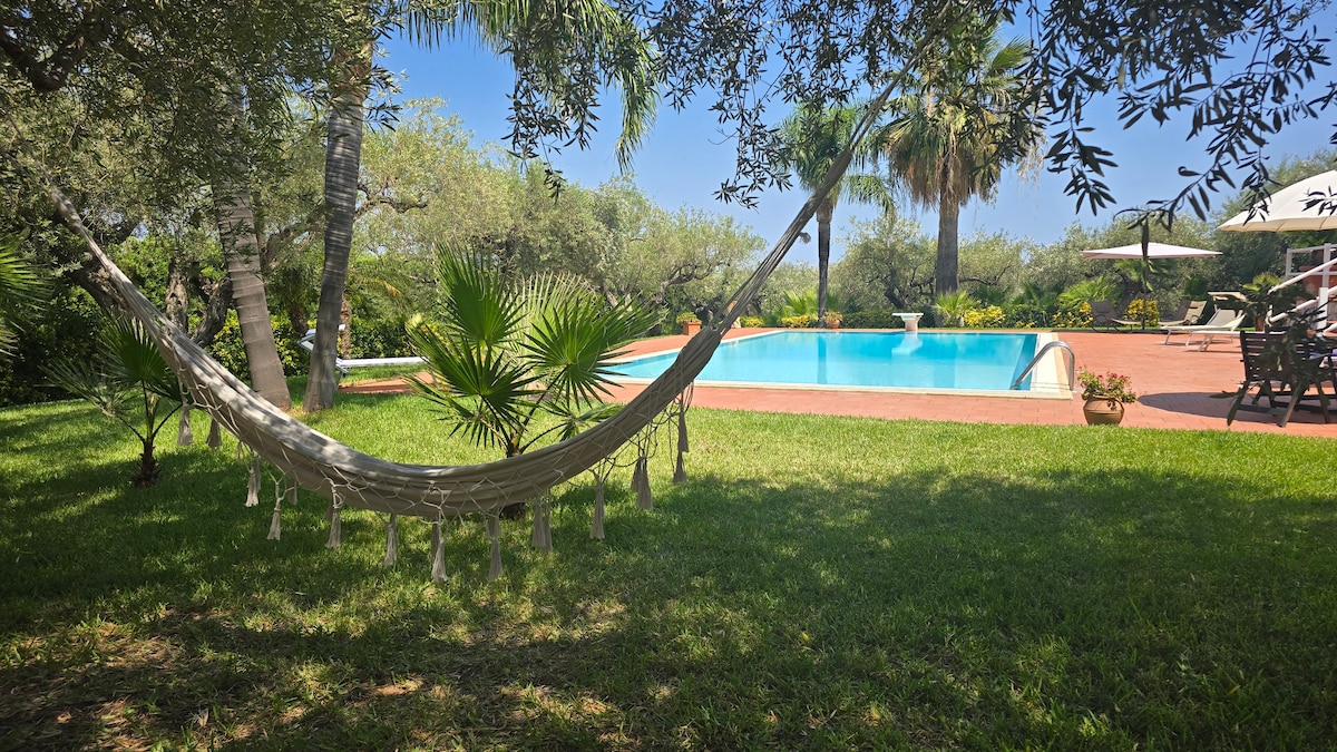 A hammock is gently swaying in a lush green garden, positioned near a private swimming pool. Surrounding palm trees and flowering plants enhance the tranquil setting, while sun loungers and umbrellas offer comfortable seating options for relaxation by the water.