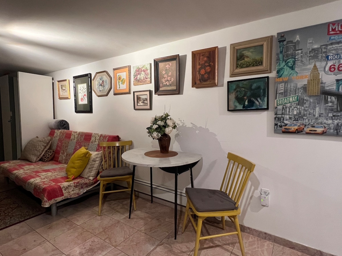 A cozy sitting area is displayed, featuring a quilted sofa with colorful patterns. A round table with two yellow chairs is positioned nearby, surrounded by a collection of framed artwork on the wall. Soft lighting creates a comfortable atmosphere.
