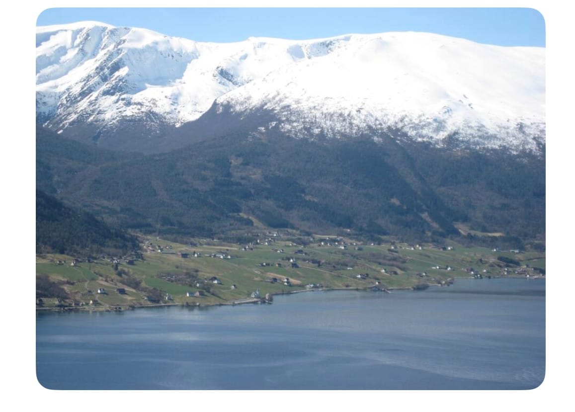 The expansive view showcases snow-capped mountains rising above a tranquil lake. Green hills gently slope toward the water's edge, where small clusters of houses are nestled into the landscape. The serene reflection of the mountains can be seen on the lake's surface.