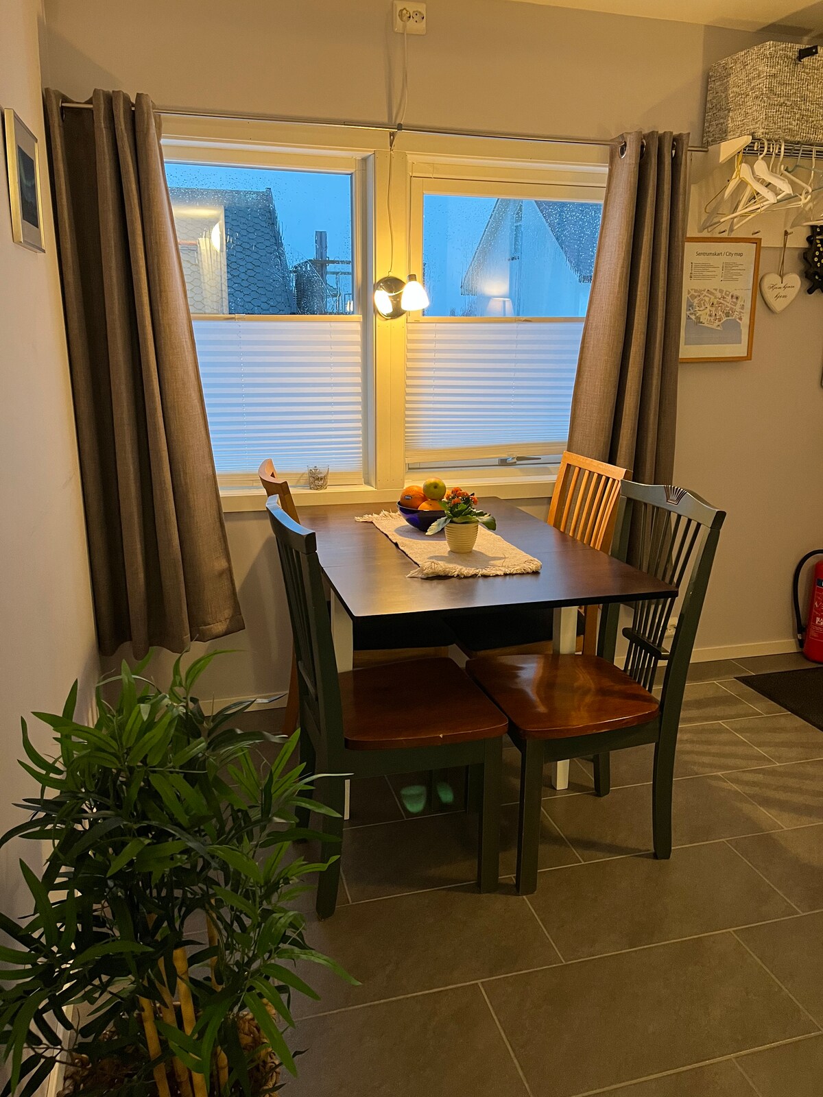 A dining area is seen, featuring a rectangular wooden table surrounded by four chairs. Sunlight filters through two windows adorned with light curtains. A small plant adds a touch of greenery, and a simple table setting with fruits is displayed at the center.
