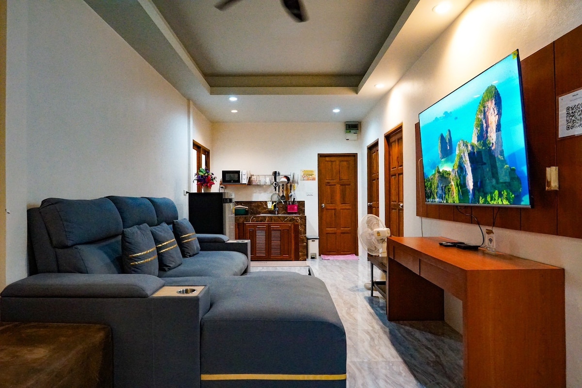 A spacious living area features a modern sectional sofa in shades of blue and a wooden entertainment center housing a large flat-screen TV. A kitchenette is visible in the background, equipped with essential appliances, while warm lighting enhances the inviting ambiance.