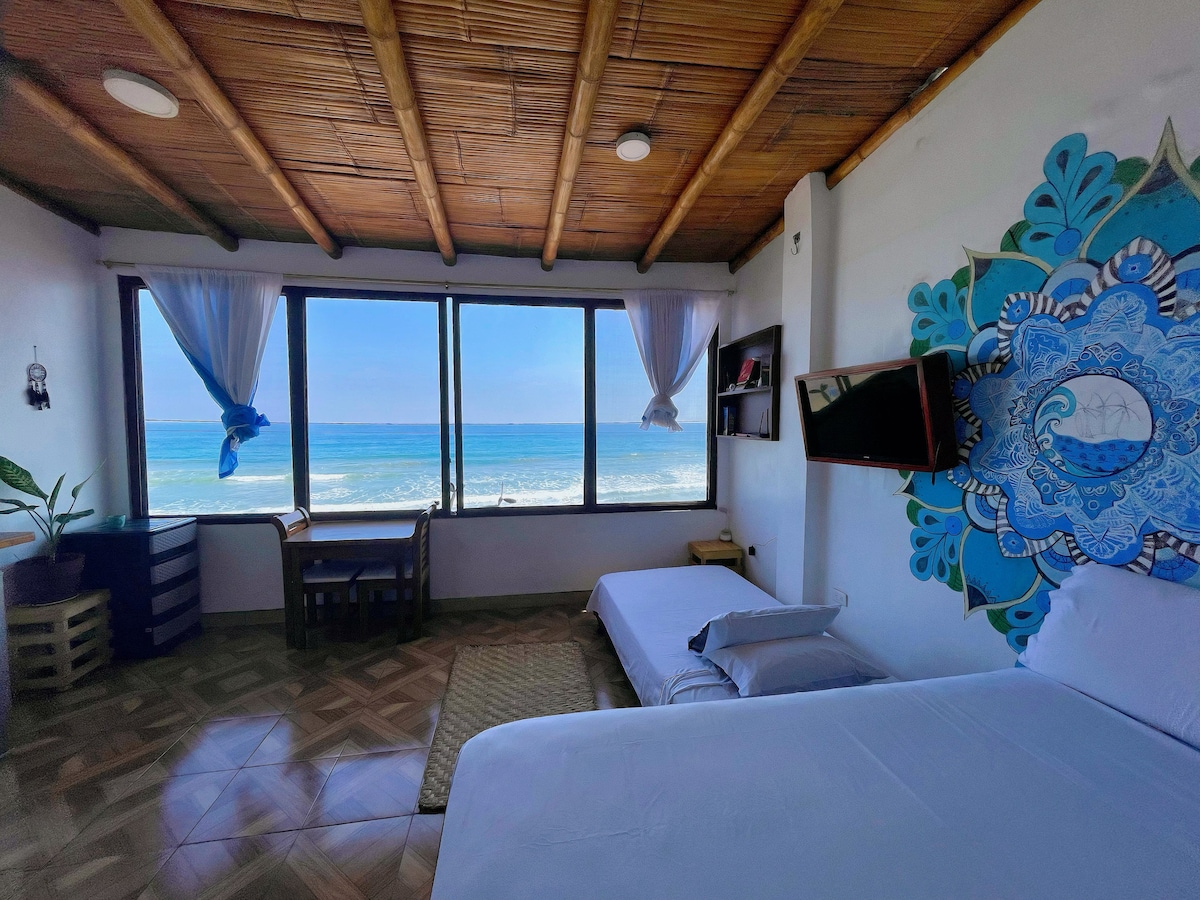 A panoramic studio features large windows offering uninterrupted ocean views. A spacious bed and a single bed are present, alongside a desk and chair. Decor includes a vibrant wall mural, and natural light fills the room, reflected by the sandy tones of the floor.