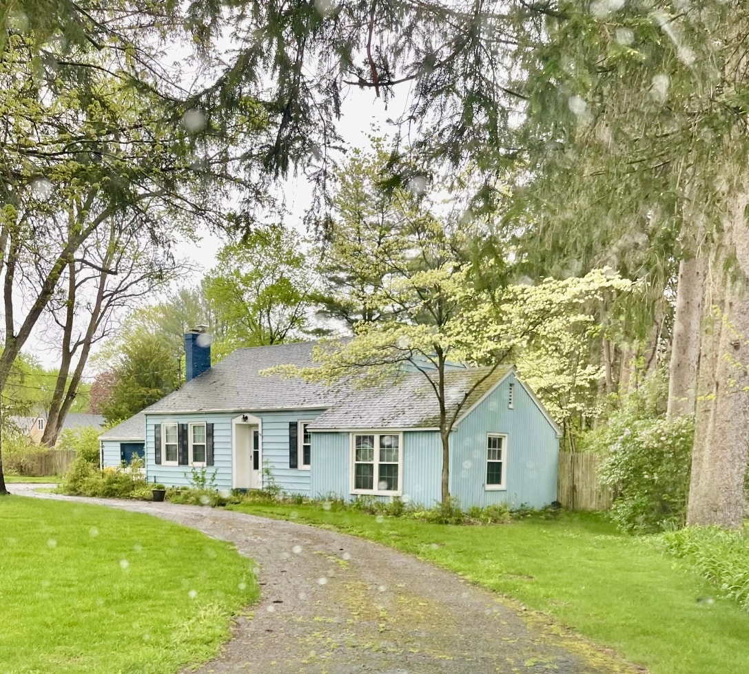 The charming blue exterior of the house is framed by lush greenery and mature trees. A gravel pathway leads to the front door, while a well-maintained lawn offers a spacious and inviting setting. Windows reflect the soft natural light filtering through the surrounding foliage.