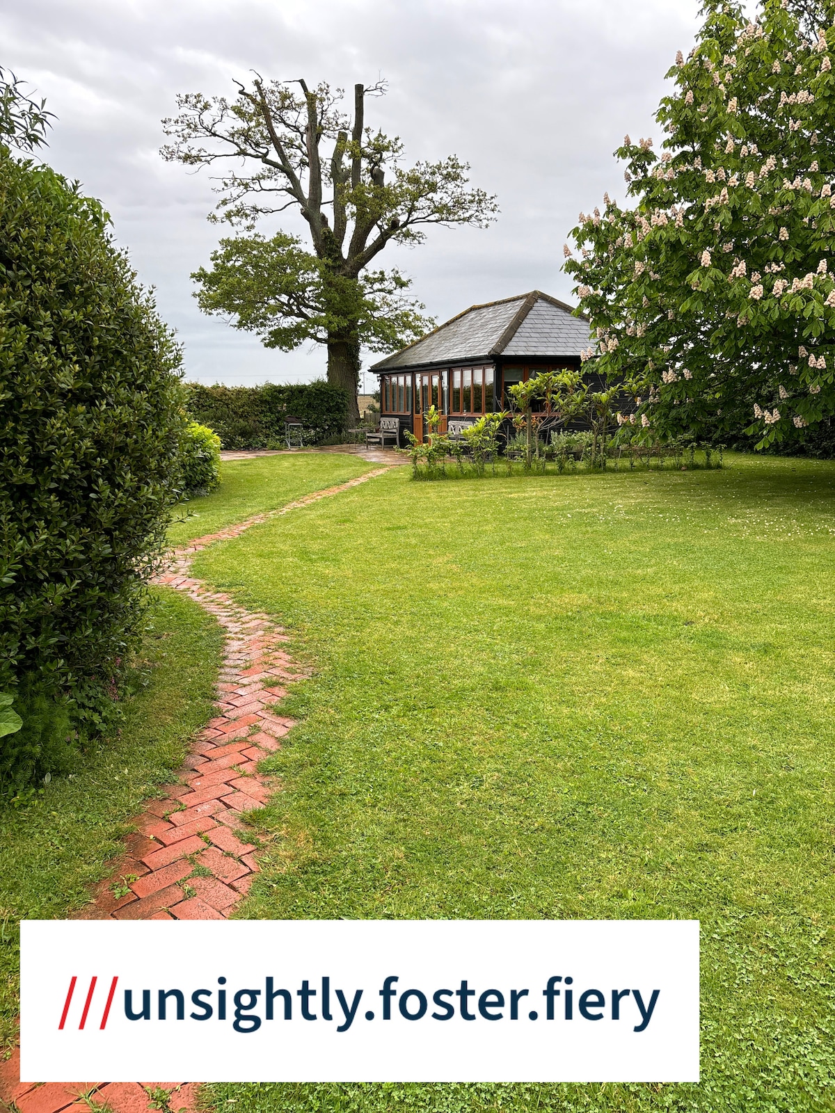 A peaceful garden path made of brick leads to the cottage, surrounded by greenery. Lush grass is present, with a large tree visible nearby. The cottage is partially obscured by plants, and the overall setting conveys a tranquil, natural environment.