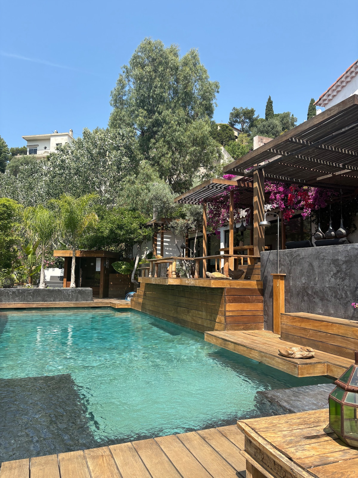 An inviting outdoor area is featured, with a clear swimming pool surrounded by wooden decking. Lush greenery and vibrant flowers are visible, enhancing the natural surroundings. A shaded dining space is positioned above the pool, creating a relaxed atmosphere.