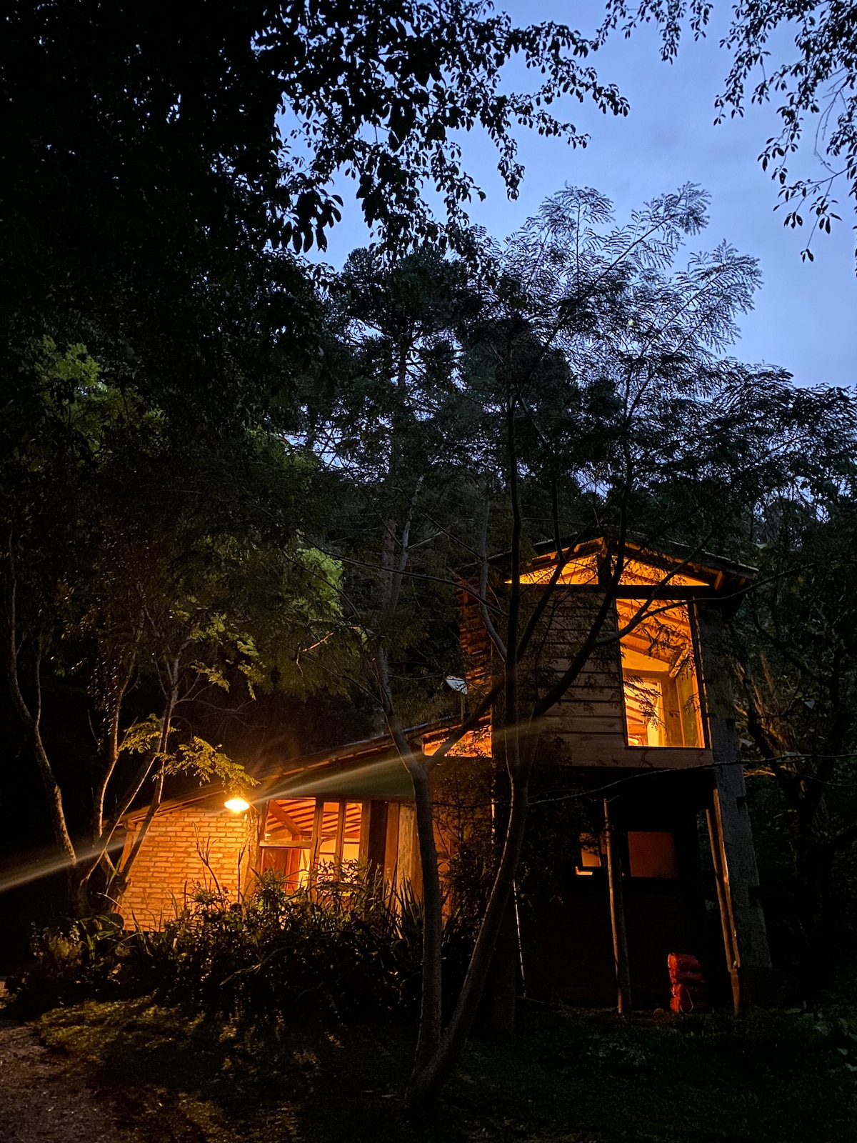 A rustic chalet is illuminated by warm light, surrounded by lush green trees. The structure features a wooden exterior, with highlights of natural elements. Soft evening light casts gentle shadows, creating a serene ambiance in the tranquil outdoor setting.