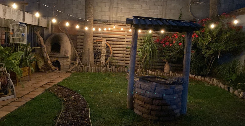 An inviting garden area is highlighted by a traditional well, surrounded by soft lighting and green grass. A rustic stone oven and wooden accents add character, while vibrant plants frame the space, creating a serene environment for relaxation or outdoor activities.