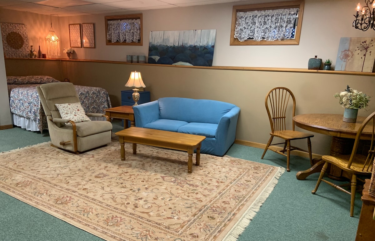 A spacious room is shown featuring a cozy sitting area with a recliner chair and a blue sofa. A wooden table with chairs is positioned nearby, along with a decorative lamp illuminating the space. A patterned rug covers the floor, complementing the inviting decor.