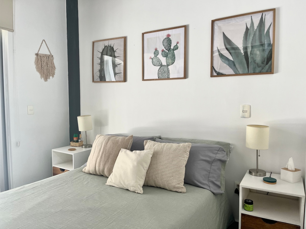 A comfortable sleeping area is showcased with a spacious bed layered in soft linens. Three framed botanical prints adorn the light-colored walls, adding a touch of nature. Two nightstands with lamps positioned on either side provide ambient lighting.
