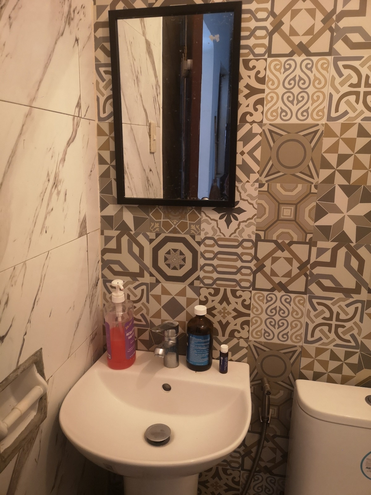 A compact bathroom features patterned tiles that add visual interest. A small sink is equipped with a wall-mounted mirror above it. Various toiletries, including a hand soap dispenser and bottles, are neatly arranged on the countertop. A modern toilet is positioned to the right of the sink.