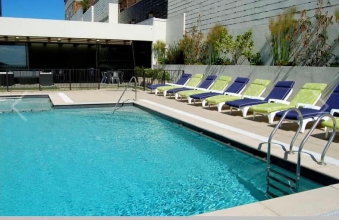 An outdoor swimming pool is framed by a terrace lined with lounge chairs featuring green cushions. The water appears clear and inviting. A shallow entry is visible, along with a backdrop of a modern building and a landscaped area, enhancing the outdoor space.