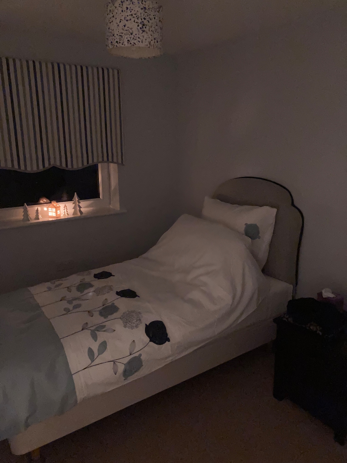 A softly lit bedroom features a single bed with a light-colored floral duvet. A small nightstand is positioned beside the bed. A decorative window treatment complements the window, where a cozy light from a decorative piece shines through.