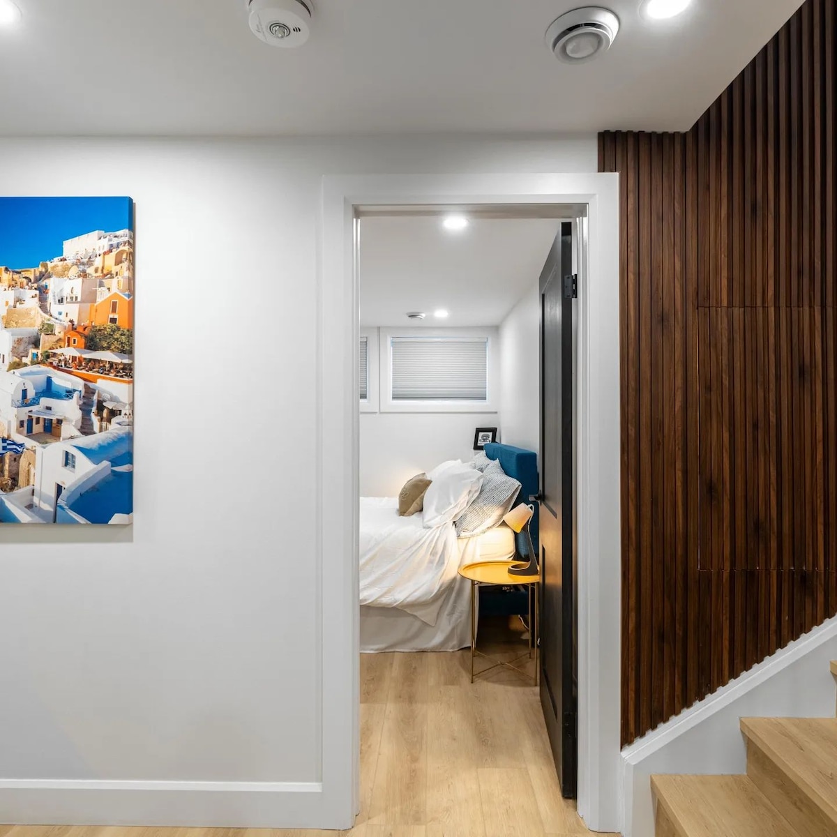A doorway leads into a cozy bedroom featuring a spacious bed with light bedding. A small nightstand is positioned next to the bed, and natural light enters through a window. The wall contrasts a wooden texture with a decorative wall art piece.