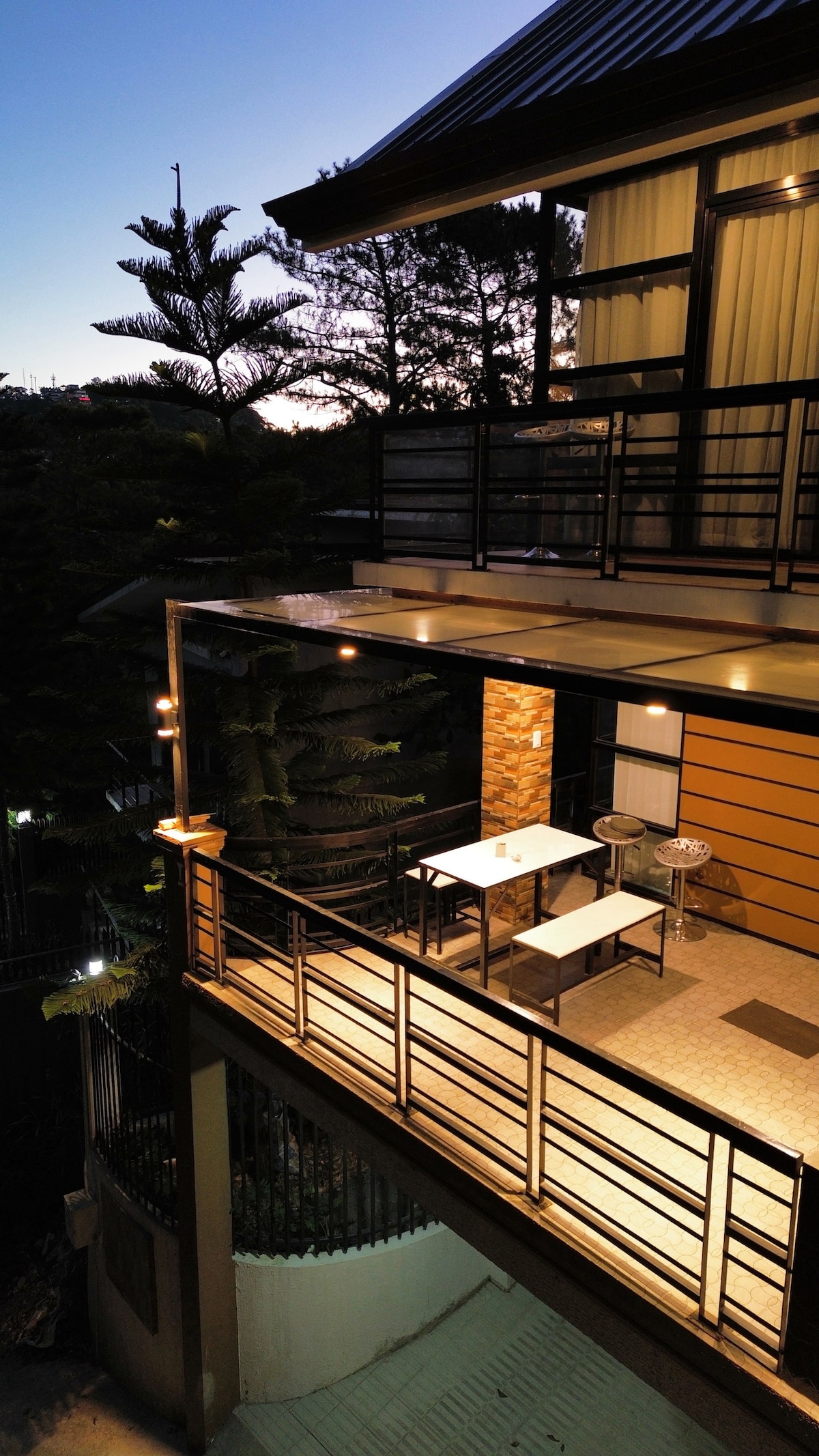 An outdoor balcony is shown with a modern design, featuring a small table and chairs. Soft lighting enhances the space, while tall trees and a view of the surrounding area are visible in the background as dusk approaches.