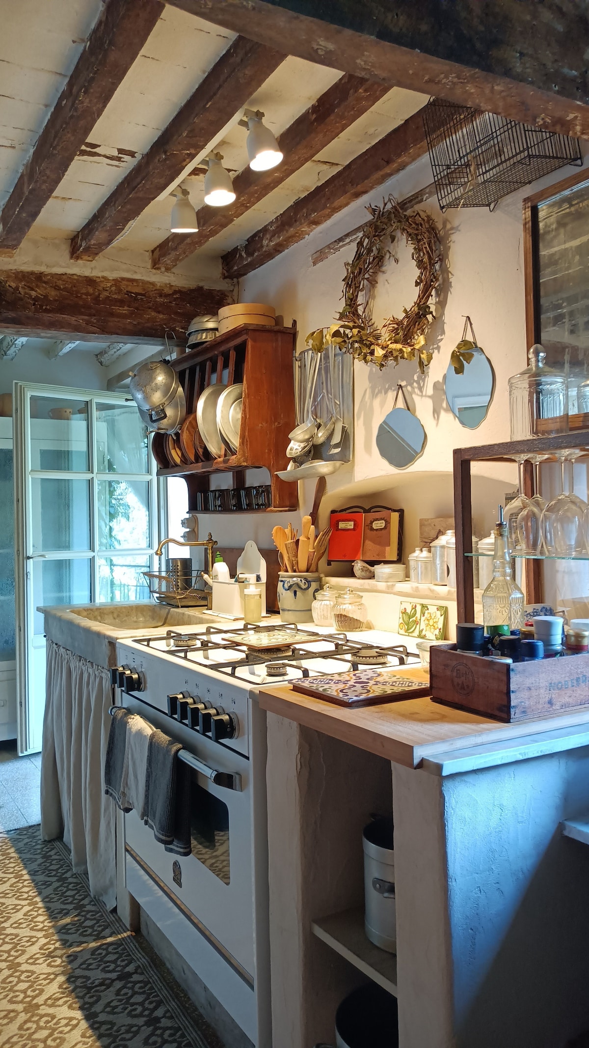A well-equipped kitchen is presented with a rustic wooden ceiling and various utensils neatly arranged on the countertops. Shelves display plates, jars, and decorative items, while natural light filters through a nearby window, creating a cozy yet functional cooking space.