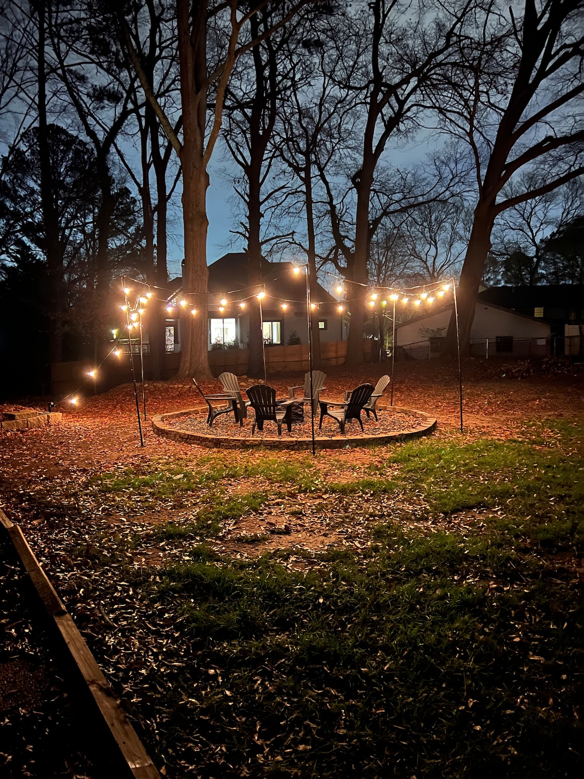 A large backyard is illuminated by string lights, creating a cozy gathering space with several outdoor chairs arranged around a fire pit. Leaf-covered ground adds a natural touch, while trees frame the area, offering some privacy.