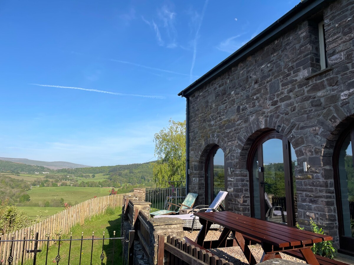The exterior of the studio showcases a stone facade with large arched windows, offering expansive views of the surrounding greenery and hills. The patio includes a wooden dining table and sun loungers, all set against a clear blue sky.