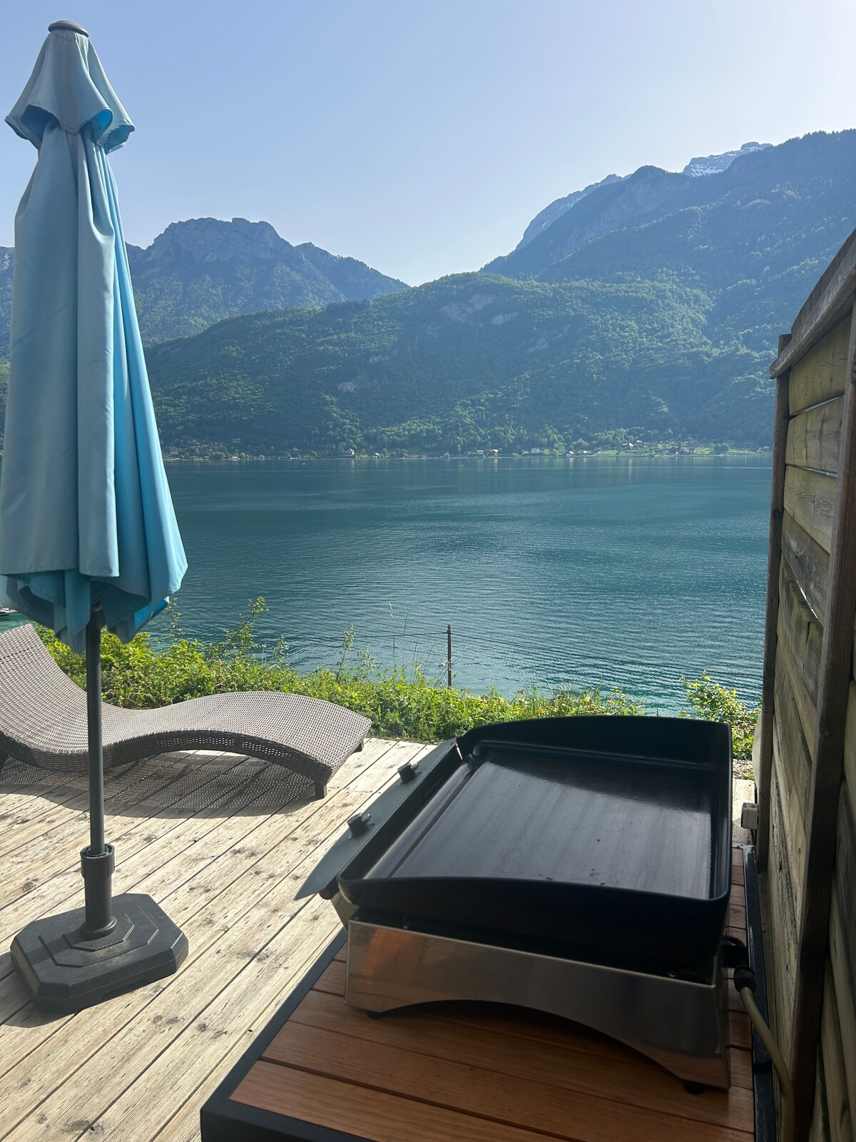 A terrace view showcases a grill on a wooden deck, accompanied by a blue umbrella. In the background, mountains rise over the tranquil waters of Lake Annecy, reflecting the clear sky.