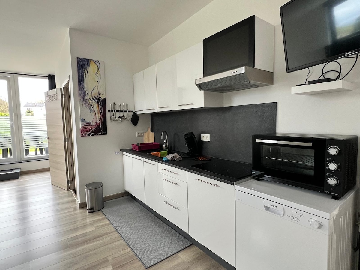 A modern kitchen is equipped with white cabinetry, a black countertop, and stainless-steel appliances. Notable features include an oven, microwave, and dishwasher. A small trash can is present, along with a mat on the floor. Natural light enters through a large window, creating a bright environment.