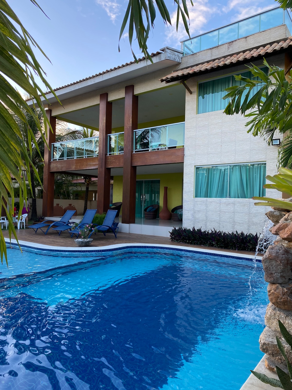 A spacious house is depicted adjacent to a clear blue pool, with lounge chairs arranged nearby. Lush greenery frames the scene, while large glass doors and windows illuminate the interior. The upper balcony offers a vantage point overlooking the pool area.