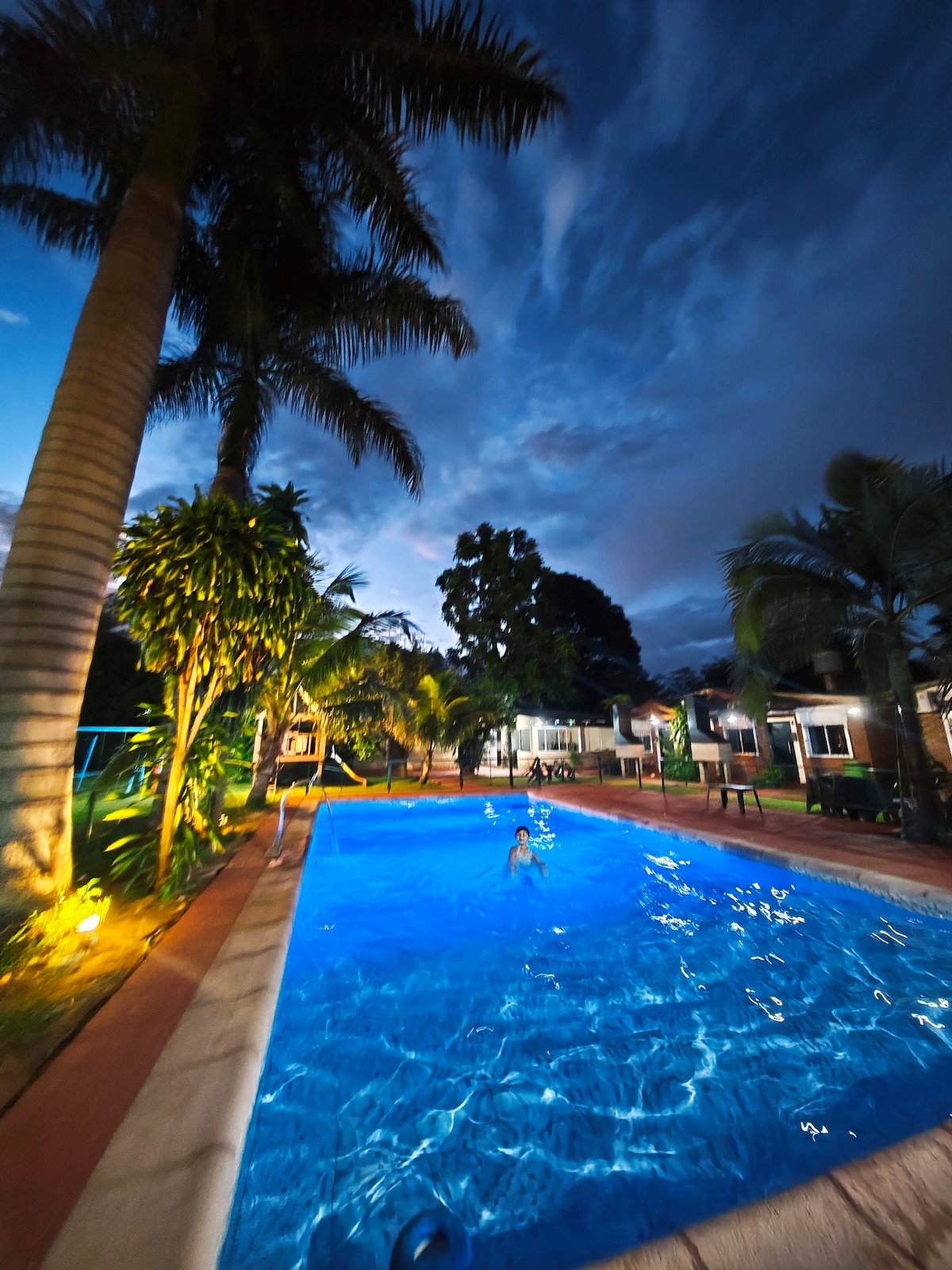 An outdoor pool glows under the evening sky, surrounded by lush palm trees and vibrant greenery. Soft lighting highlights the area, creating a serene atmosphere. Cabanas and seating are visible in the background, enhancing the inviting setting for relaxation and leisure.