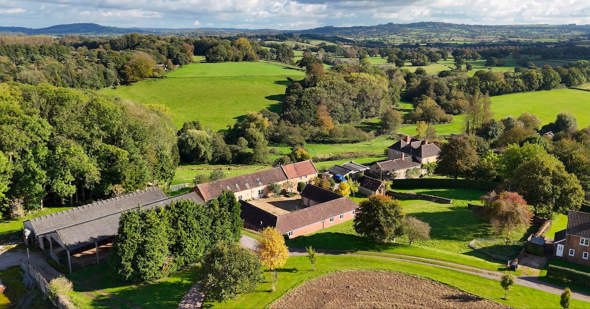 An aerial view of Higher Easthams Farm is shown, revealing a collection of buildings nestled amid lush green fields and wooded areas. The landscape includes rolling hills in the distance under a partly cloudy sky, emphasizing the spacious and serene rural setting.