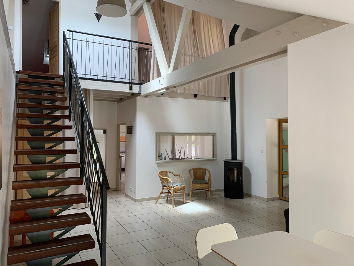 An inviting living space features high ceilings supported by wooden beams. A staircase with a black railing leads to the upper level. Two rattan chairs are placed near a window, and a stove is located against the wall, complementing the open layout.