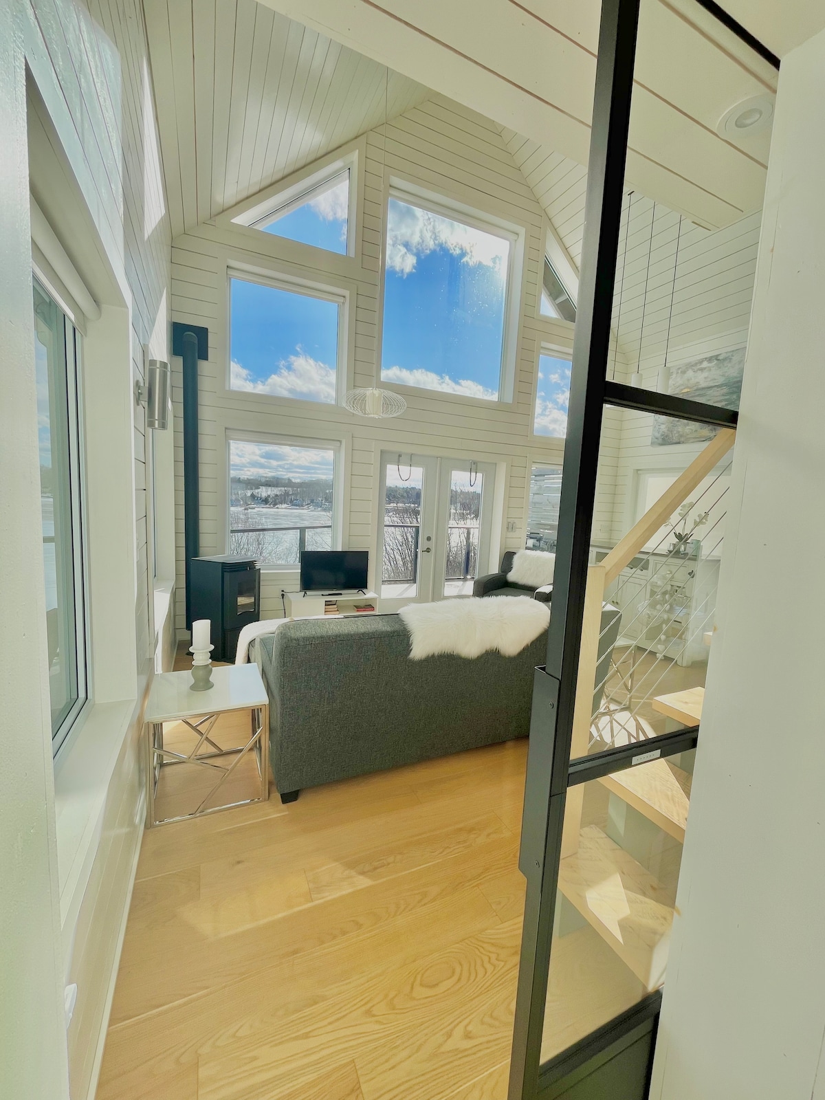 Bright, airy living space is illuminated by large windows offering expansive views of the ocean and surrounding landscape. A comfortable sofa is paired with a chic side table, while a sleek fireplace adds a touch of warmth. Access to a wooden staircase leads to the loft above.