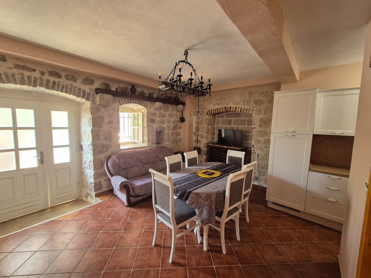 An inviting dining area features a rectangular table surrounded by chairs, with a stone wall providing texture. A comfortable sofa sits nearby, and a rustic chandelier hangs from the ceiling. Natural light enters through French doors, enhancing the room's warm ambiance.