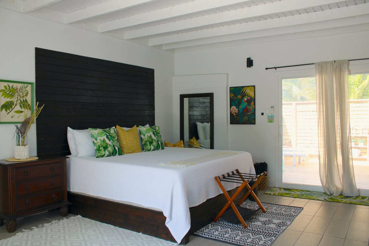 A spacious sleeping area features high ceilings with white beams and bright white walls. A large bed is accompanied by decorative pillows in vibrant colors. Nearby, sliding glass doors provide access to a private deck, enhancing the natural light within the room.