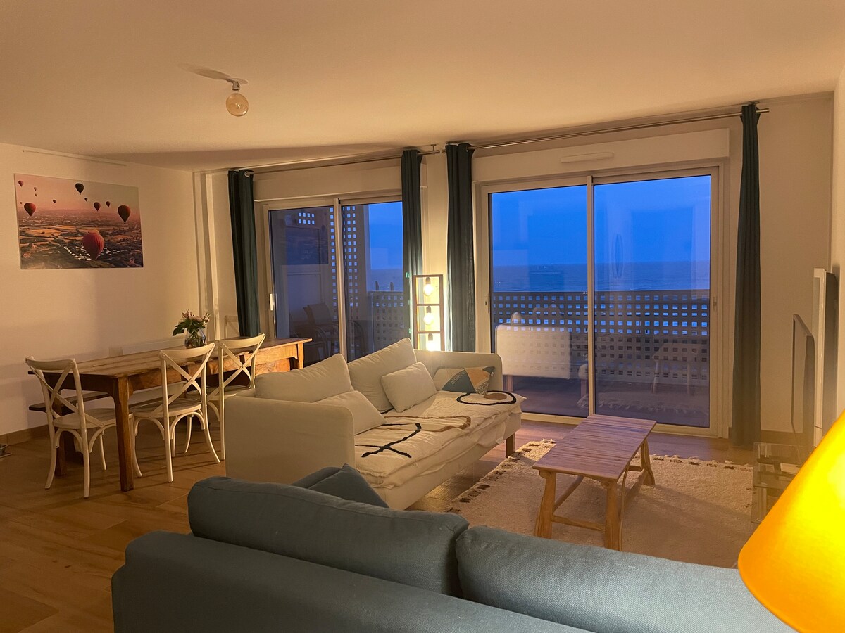 A spacious living area features large glass doors that open to a terrace overlooking the ocean. Two comfortable sofas are arranged around a wooden coffee table. A dining table set for four is positioned nearby, complemented by a warm glow from a standing lamp. Soft lighting enhances the evening ambiance.