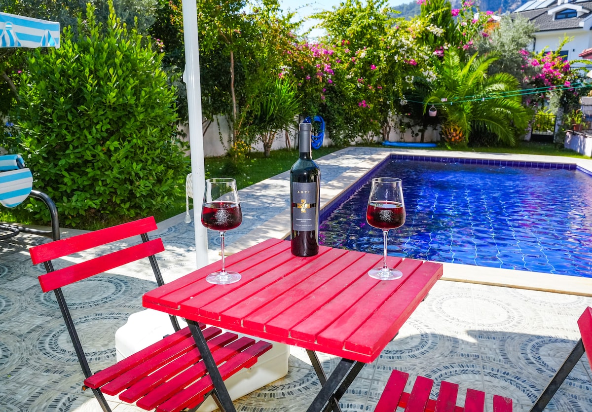A small outdoor dining area is featured, with a red table set for two glasses of wine. A bottle rests in the center. Behind the table, a blue tiled pool glimmers under sunlight, surrounded by lush greenery and flowering plants.