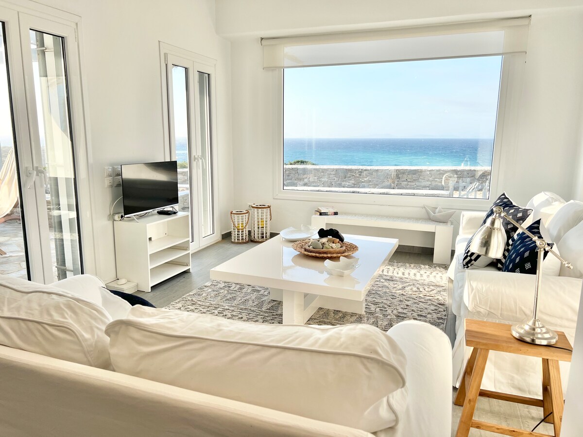 A bright living space features a large window offering unobstructed views of the Aegean Sea. Comfortable white sofas are arranged around a central coffee table, with a small wooden side table beside one sofa. The decor is minimalistic, complemented by a patterned area rug underfoot.