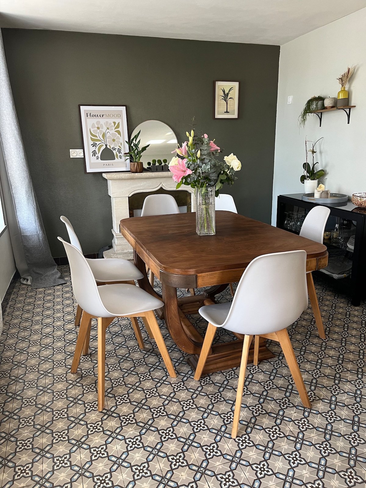 A dining area features a wooden table surrounded by four modern chairs. A vase of fresh flowers centers the table. A large round mirror and wall art are displayed against a dark green wall, while patterned tiles add visual interest to the floor.