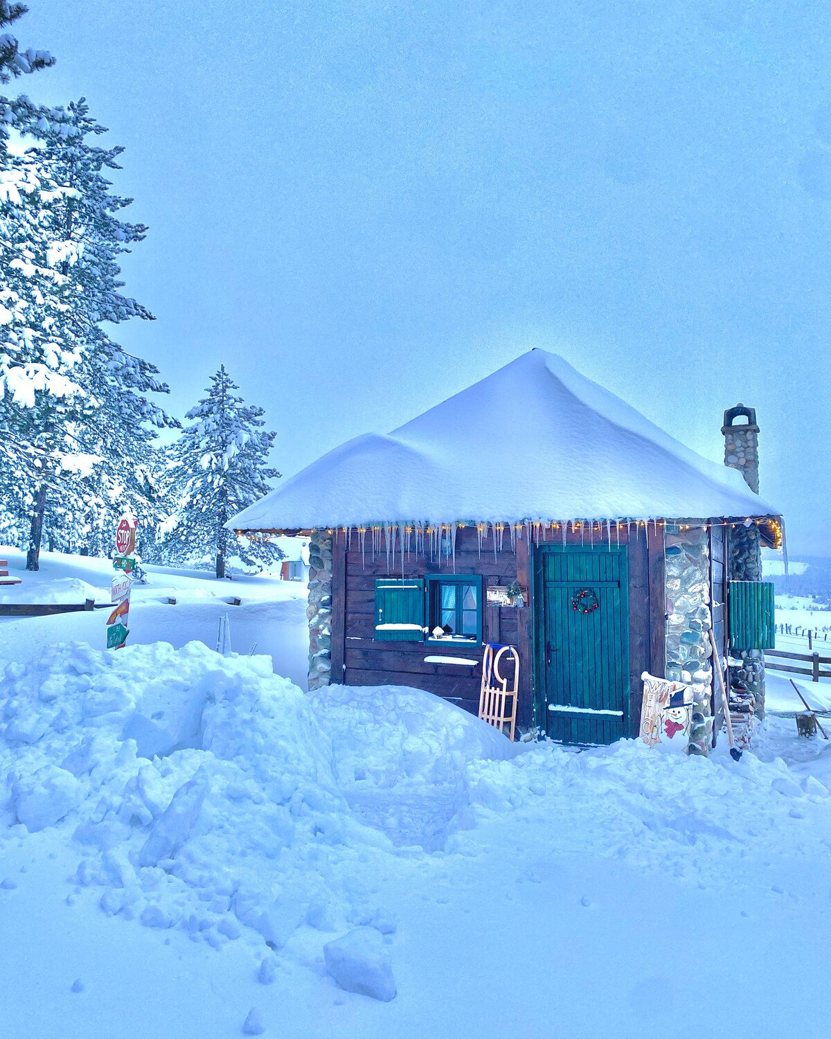 A charming cabin is surrounded by a blanket of snow, with a sloped roof capped in white frost. Icicles hang from the eaves, and a green door adds a pop of color against the winter landscape. Nearby trees are dusted with snow, enhancing the serene setting.