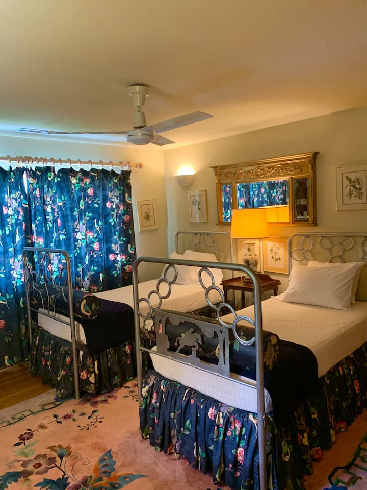 This charming guest room features a maximalist aesthetic, blending vintage elegance with bold, botanical patterns. Two unique, custom wrought-iron twin beds—one notably adorned with a silver bronc rider centerpiece—providing cozy and artistic warmth