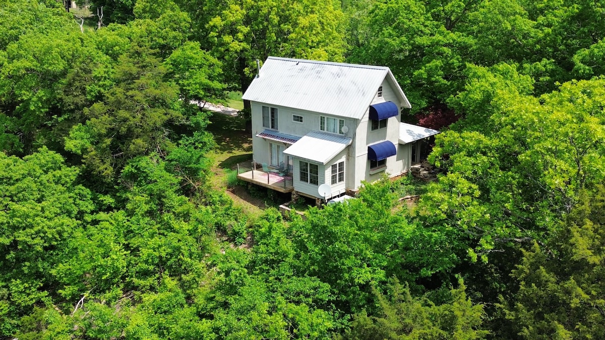 The cottage is surrounded by lush greenery, featuring a metal roof and multiple windows. The deck extends from the front, offering a view of the surrounding oak trees. The serene setting emphasizes the natural beauty of the Ozark mountains.
