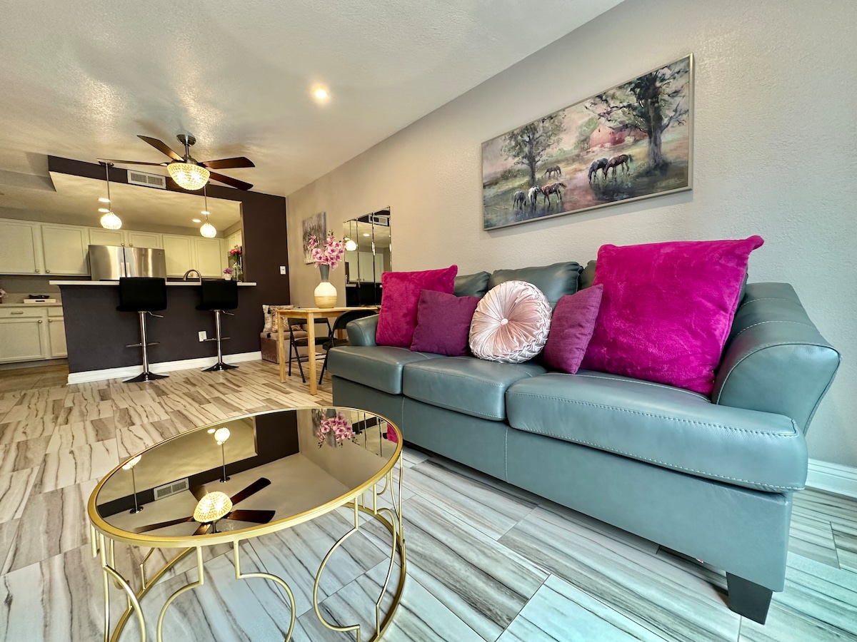 A leather couch featuring decorative pillows is positioned in the living area. A round coffee table with a reflective surface is nearby. A dining table is slightly visible in the background, complementing the open layout. Natural light brightens the space, accentuating the modern decor.