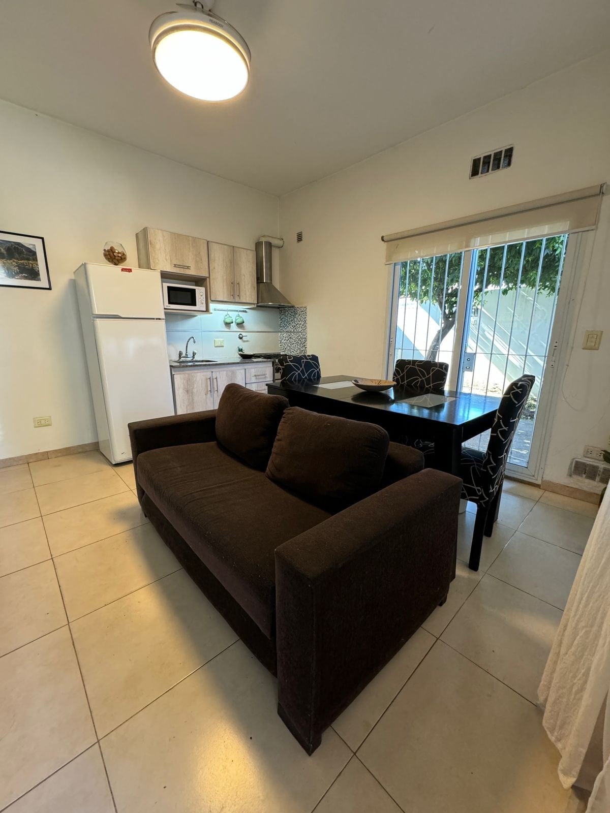 A comfortable living area features a dark sofa and a dining table with elegant chairs. The kitchenette, equipped with essential appliances, is visible in the background. Large glass doors open to the outside, allowing natural light to illuminate the space.