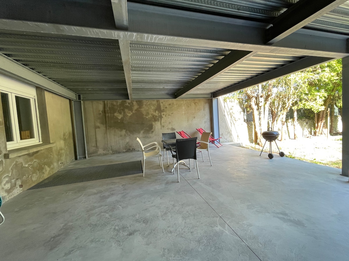A covered terrace features a spacious, concrete surface. Several dining chairs surround a small table, complemented by two lounge chairs in the background. A charcoal barbecue stands to one side, with greenery visible beyond the terrace, creating a peaceful outdoor setting.