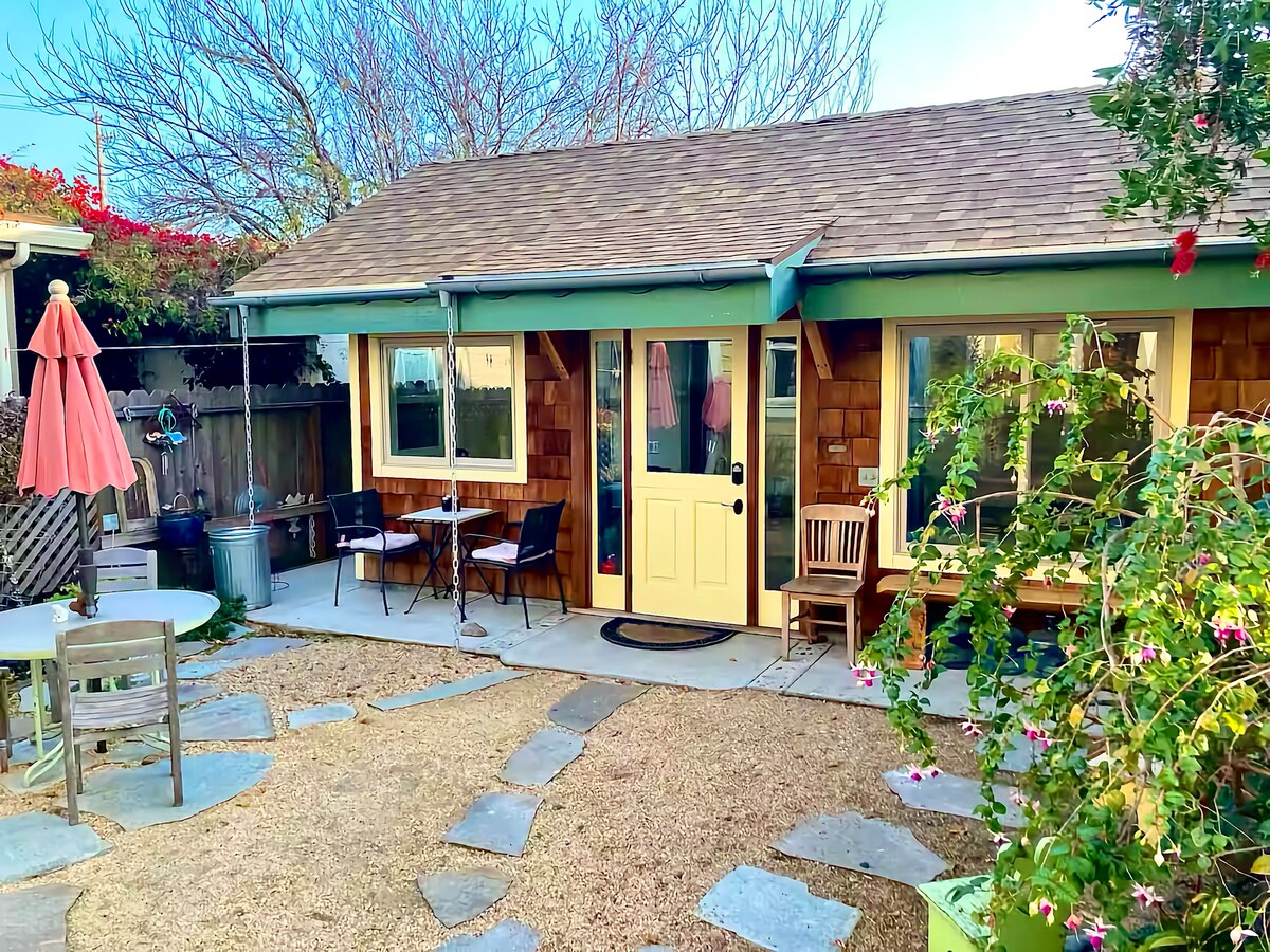 A charming cottage is presented with a wooden exterior and a bright yellow door. Outdoor seating is provided at small tables and chairs, complemented by a vibrant umbrella. A gravel path leads through the garden, with lush greenery surrounding the entrance.