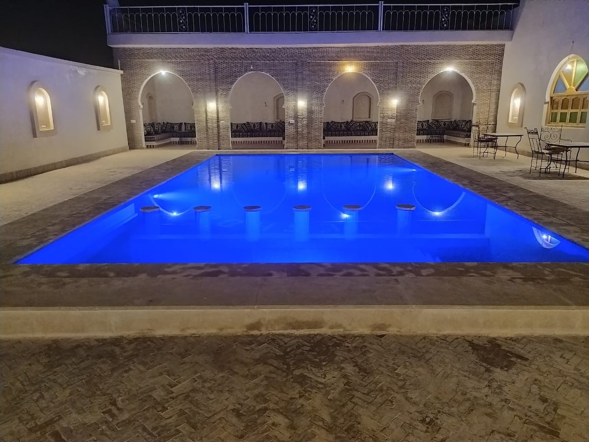A rectangular swimming pool is illuminated with a soft blue light, surrounded by textured stone flooring. Arched doorways and a seating area with black furniture are visible in the background, contributing to a relaxing outdoor atmosphere.