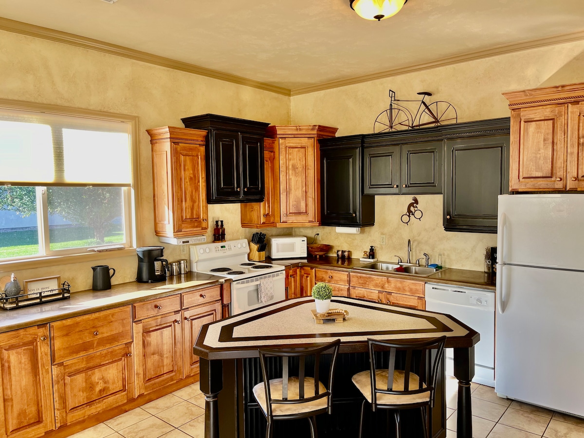A spacious kitchen is highlighted by wooden cabinetry and neutral-toned walls. The kitchen features an island with seating, essential appliances including a fridge and dishwasher, and ample counter space for meal preparation. Natural light enters through a large window, creating a warm and functional area.