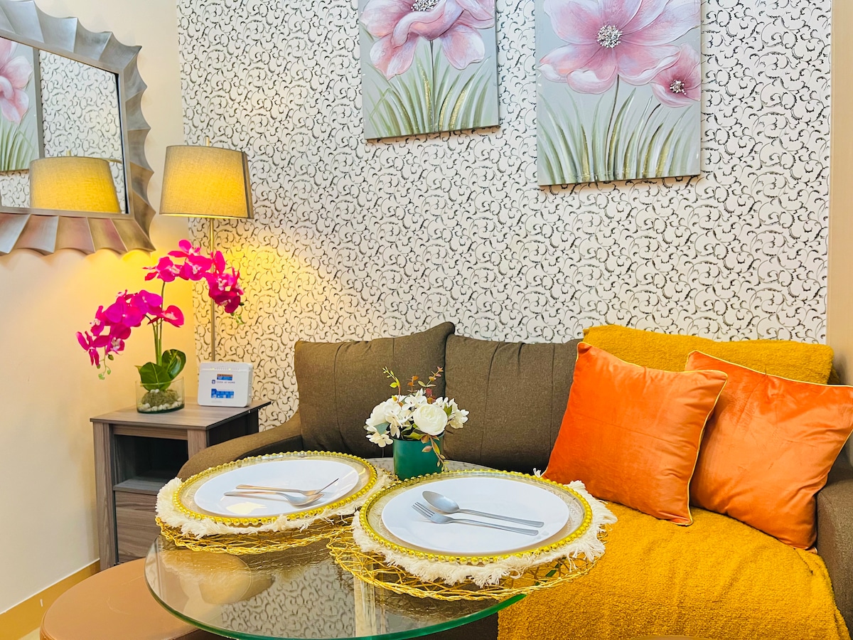 A cozy seating area is highlighted by a four-seater sofa adorned with orange cushions. A glass coffee table is set with place settings, accompanied by a small vase of flowers. Two floral wall art pieces bring a touch of color to the wall behind.